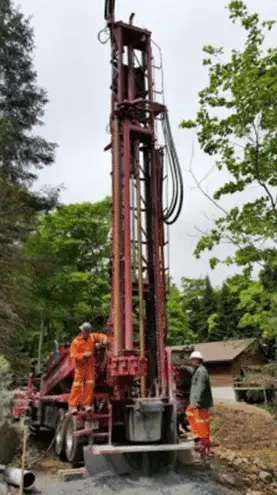 Two workers of groupe h2o in orange safety suits work around a specialized drill rig for drilling an artesian well in a forested environment, with a building visible in the background.