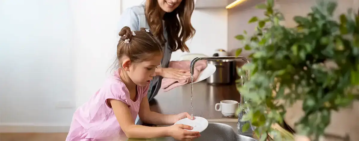 A little girl in a pink dress washing a plate under running water in a sink, assisted by her smiling mother in the background, in a modern kitchen adorned with green plants. traitement de l’eau